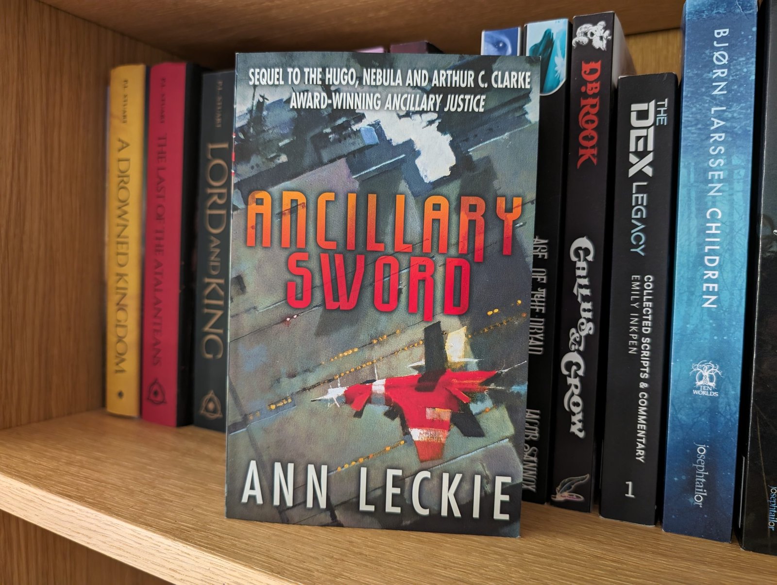 The novel Ancillary Sword by Ann Leckie. A small red spacecraft flies over the surface of a much larger green spaceship.
