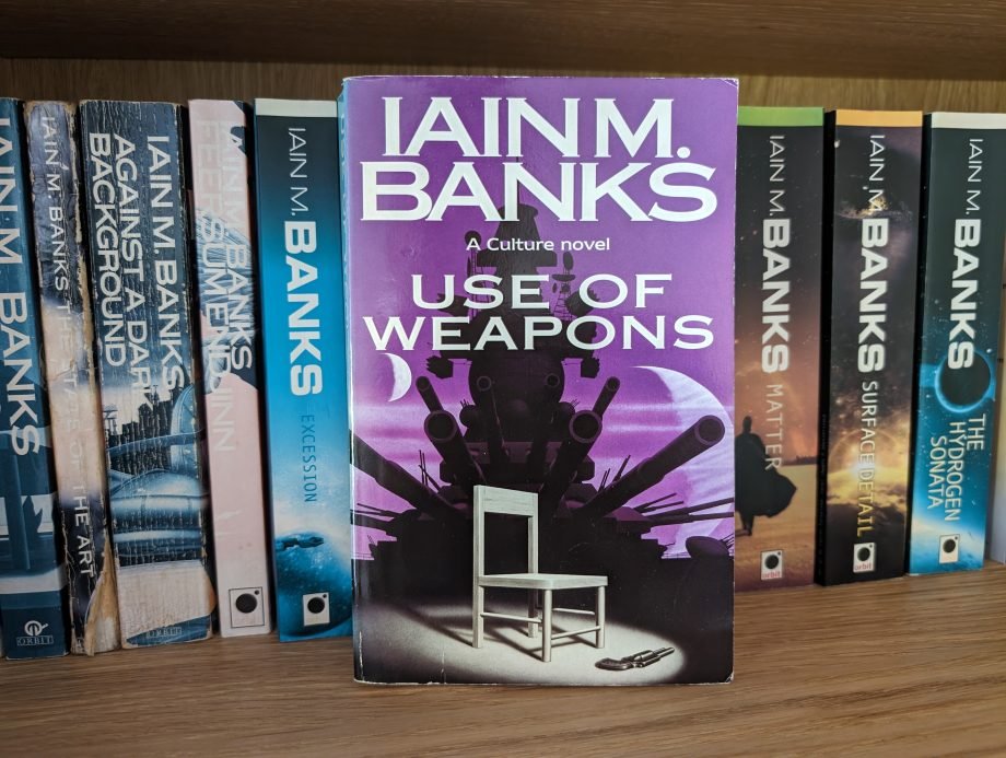 Use of Weapons by Iain M Banks The novel Use of Weapons by Iain M Banks, which features the black silhouette of a battleship. In front of the ship is a white chain and a black revolver lies on the floor in front of the chair. The book is pictured on a shelf, next to Iain M Banks other sci-fi novels.